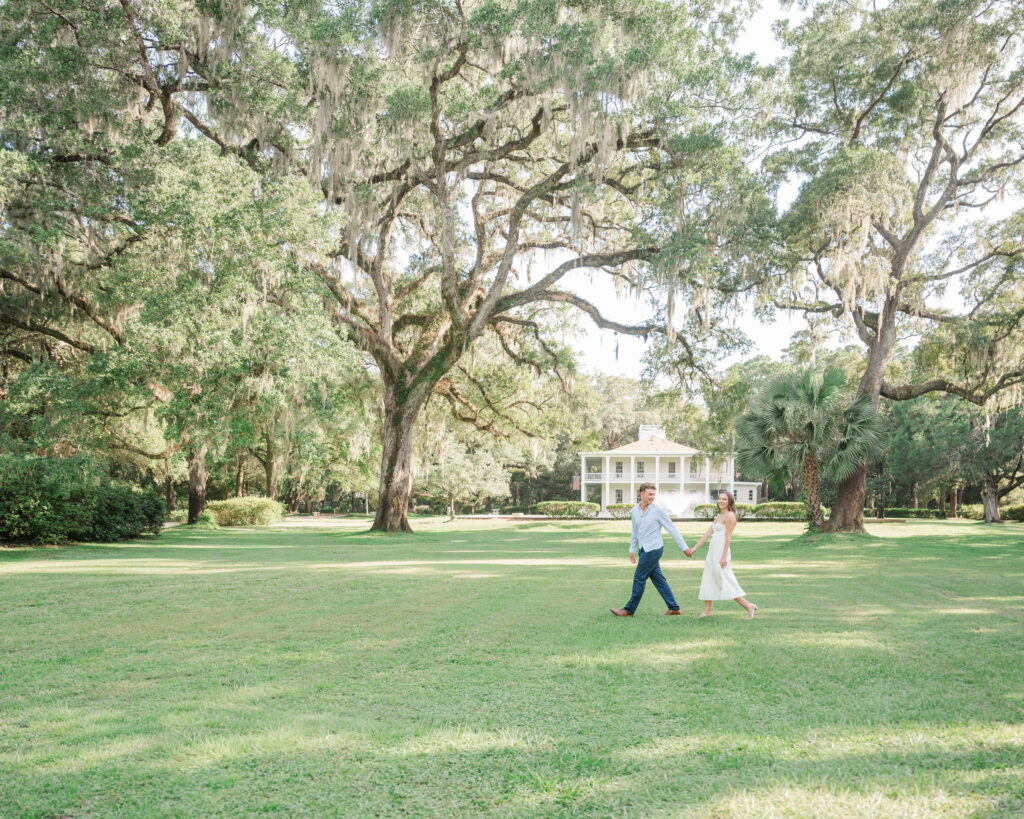 Engaged couple walking hand in hand across the expansive lawn beneath live oaks at Eden Gardens State Park in Santa Rosa Beach, Florida.